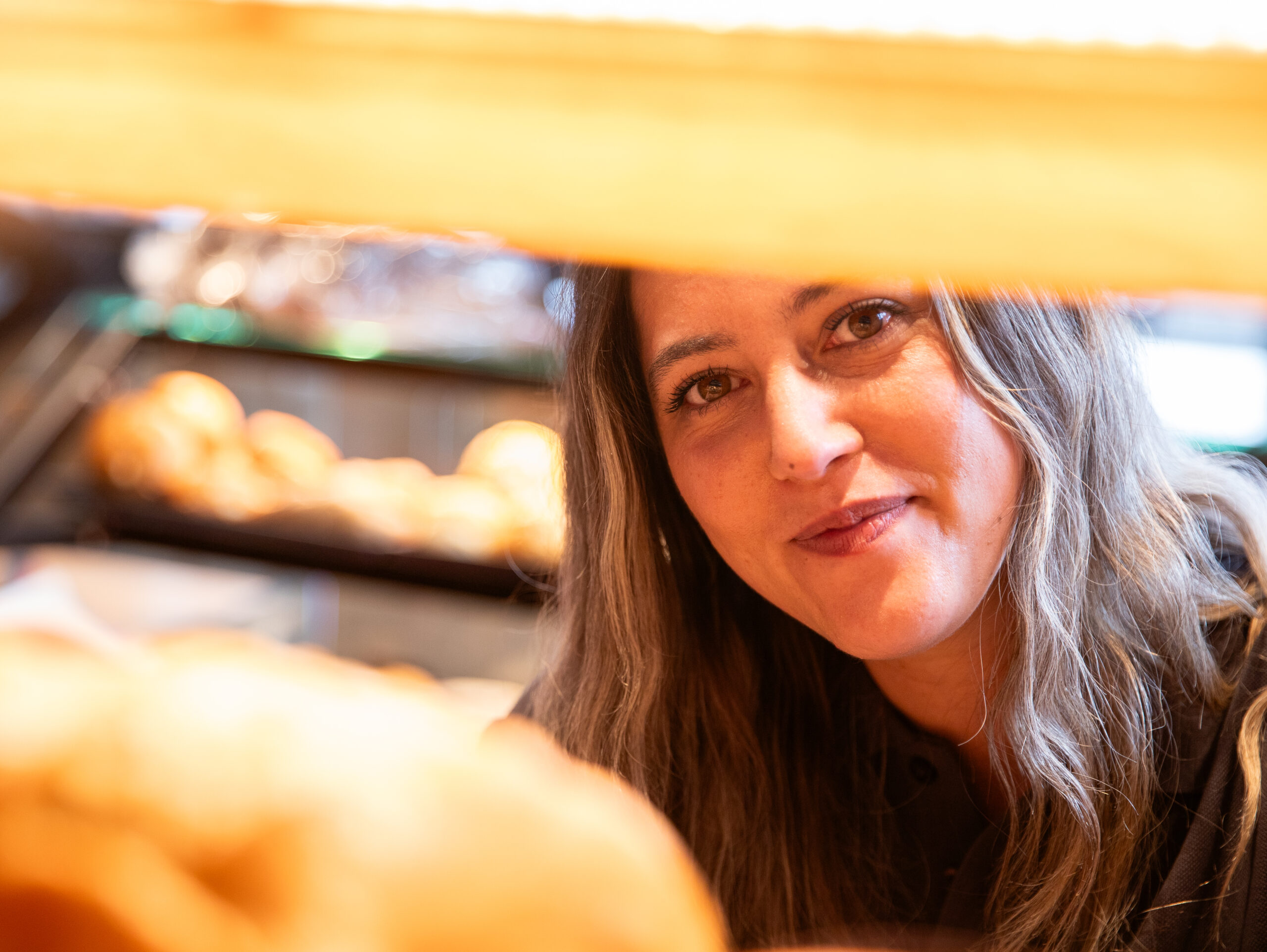 Bäckerei Fachverkäuferin, ca. 37 Jahre alt, schau lächelnd zwischen Blechen mit Brötchen hindurch die auf einem Backblechständer stehen in einer Bäckerei Filiale.