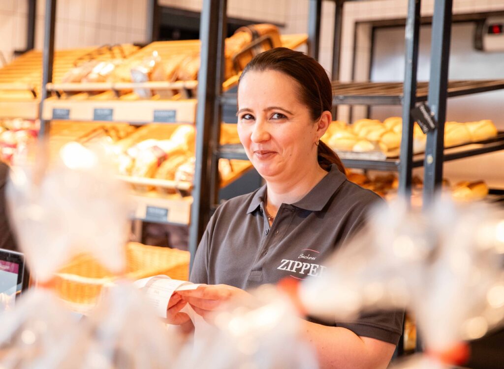 Bäckerei Fachverkäuferin, ca. 40 Jahre alt, steht in Arbeitskleidung hinter der Theke einer Bäckerei Filiale und hält Kassenbelege in beiden Händen.