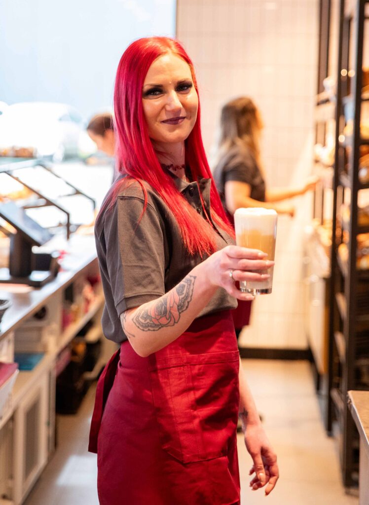 Bäckerei Fachverkäuferin, ca. 32 Jahre alt, steht in Arbeitskleidung hinter der Theke einer Bäckerei Filiale mit einem Kaltgetränk in der Hand.